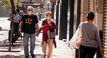 Pareja de adultos mayores con mascarilla y escudo facial caminando por una vereda de Santiago un día soleado.