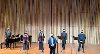Foto panorámica del escenario del Centro de Extensión Oriente, con cuatro cantantes de pie, con mascarillas y partituras. Además de un pianista y piano de cola en el fondo de la imagen y al centro el director del coro de cámara UC. Todos con ropa de presentación formal.