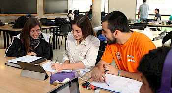 dos estudiantes mujeres y un estudiante hombre sentados alrededor de una mesa con lápices, cuadernos y tablet
