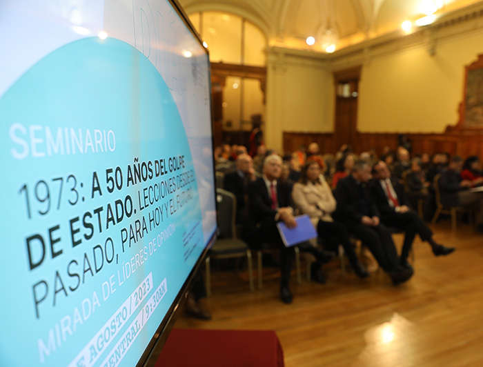 Pantalla en primer plano con la imagen del seminario y público desenfocado de fondo.