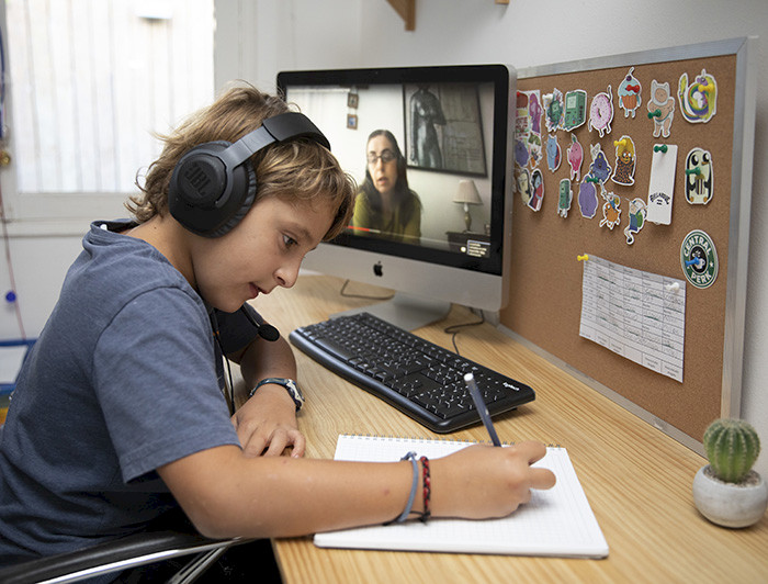 Niño escribe sobre un cuaderno usando audífonnos y junto a un computador donde se ve una mujer.