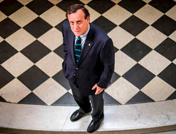 Rector Ignacio Sánchez mira hacia arriba de pie sobre un fondo de baldosas negras y blancas.